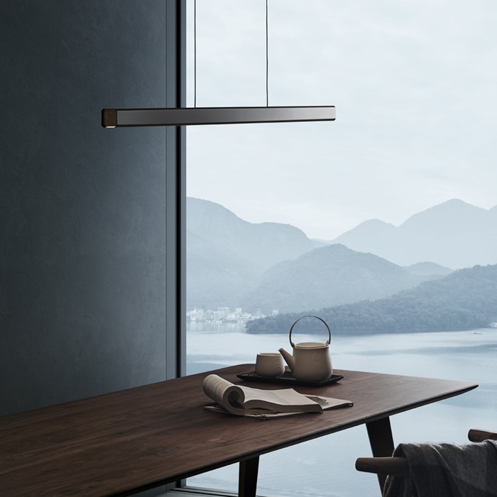 Lifestyle image of the Seed Design Mumu 120 black and walnut pendant hanging above a walnut wood table and next to a large window looking out at mountain scenery