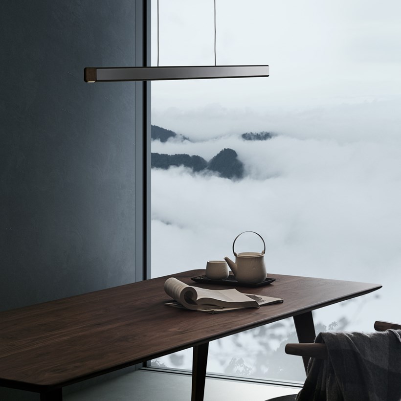 Lifestyle image of the Seed Design Mumu 120 black and walnut coloured LED pendant suspended above a walnut table, with heavy sea waves in the background