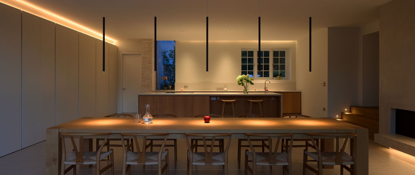 Contemporary kitchen dining room, with 5 black tubular pendants suspended over a long dining table & linear LED above the kitchen cupboards