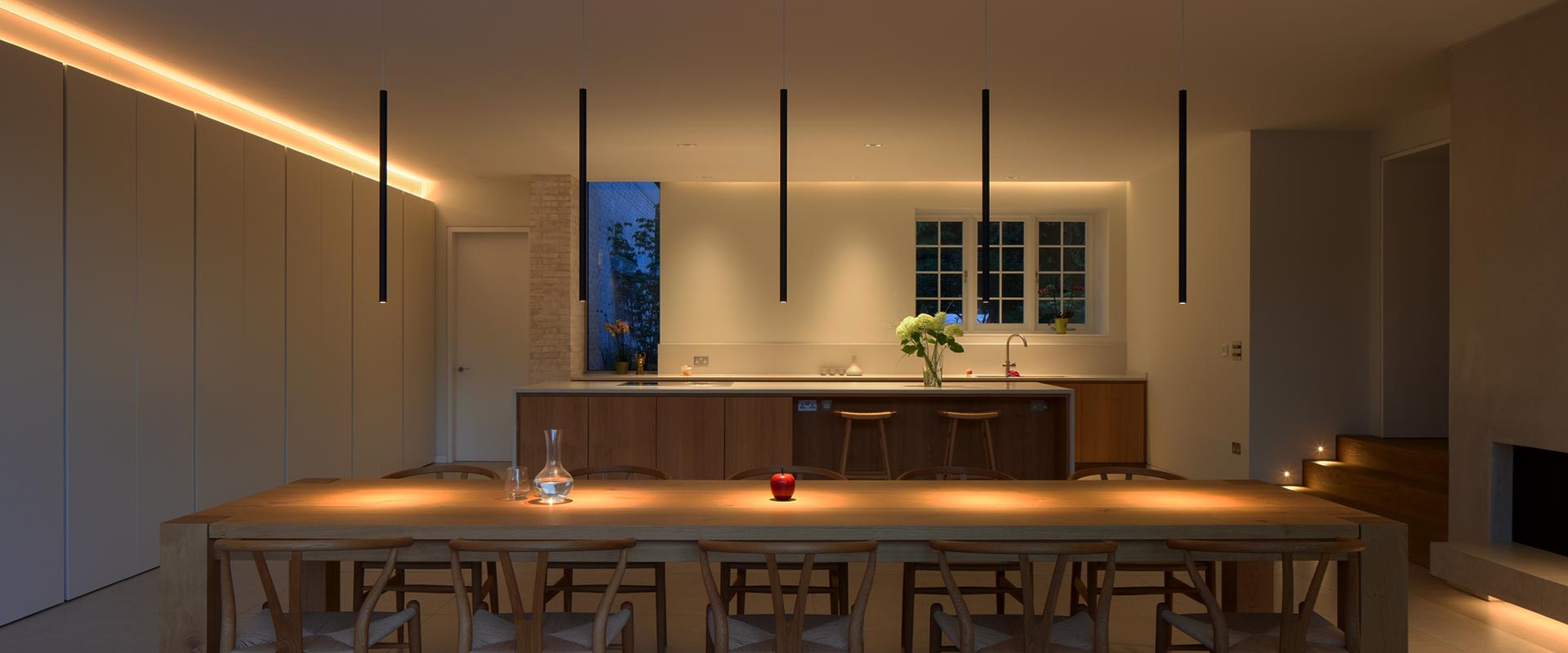 Contemporary kitchen dining room, with 5 black tubular pendants suspended over a long dining table & linear LED above the kitchen cupboards