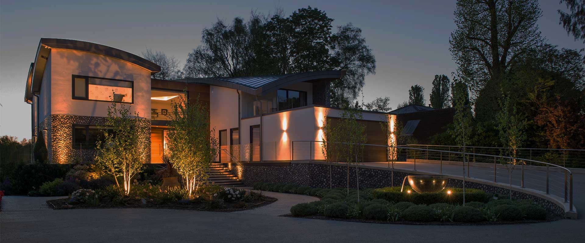 Modern house, garage and front driveway lit with up/down wall lights, with landscape lighting spike spotlights & recessed floor lights at dusk