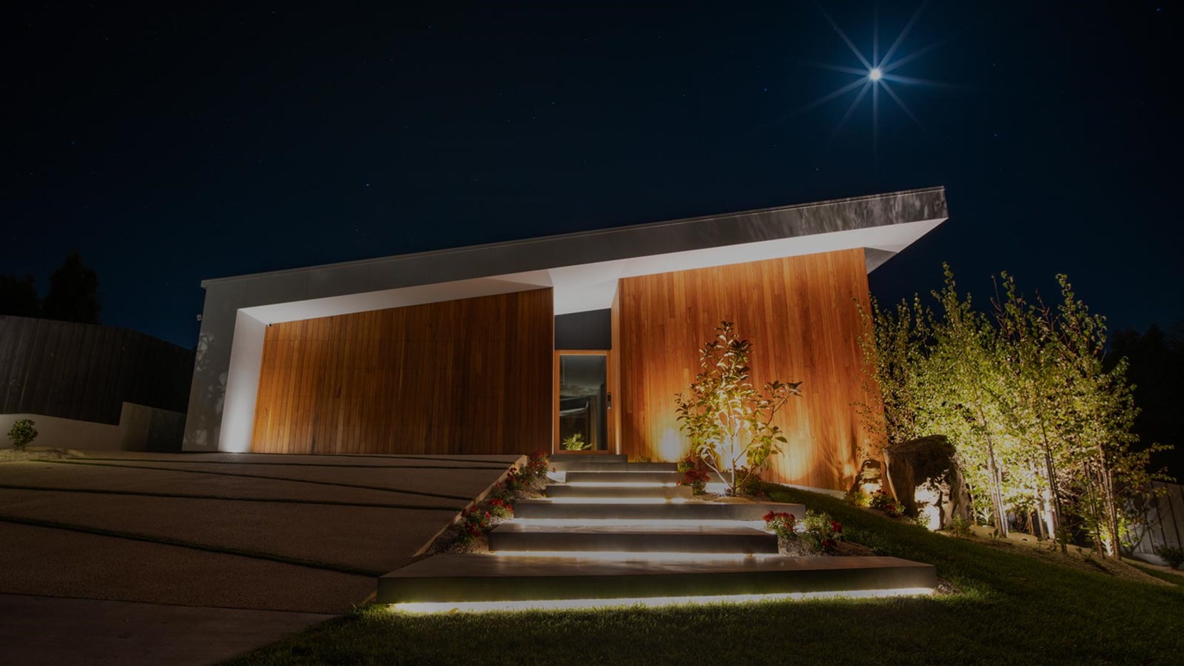 Outside of modern minimal asymmetrical house at night, lit with uplights. The steps are lit with strips of linear light