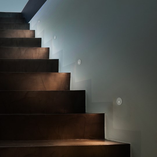 Architectural recessed round low level LED step light recessed into the wall, lighting down onto a modern set of stairs
