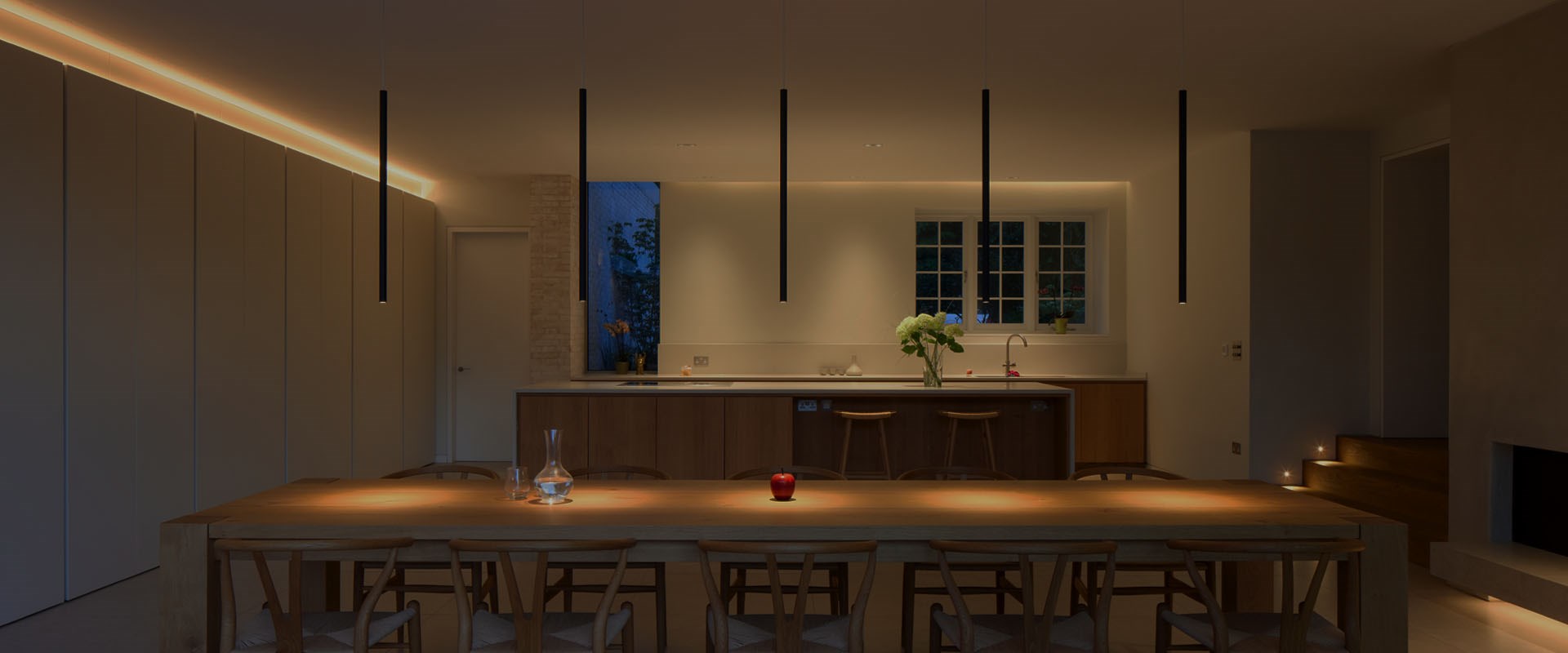 Shop: Modern kitchen diner with 5 black tubular pendants suspended over a dining table, linear LED above the kitchen cupboards, step lights in the wall